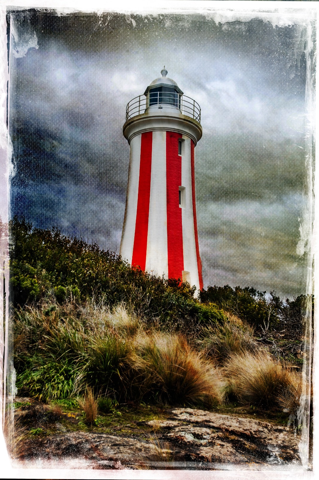 A striped lighthouse rising from coastal vegetation under a darker, storm-weighted sky.